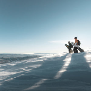Image of two people standing side by side with snowboards on a ski hill against a blue sky.